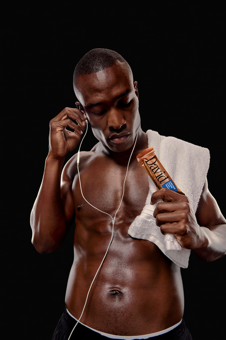 Muscular man wearing earphones and a towel over his shoulder holding a David Cookie Dough Caramel Chocolate Crunch protein bar